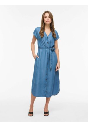 Vrouw staande, gekleed in een blauw midi-jurk met knopen en tailleband en zwarte open-toe hakken tegen een effen witte achtergrond