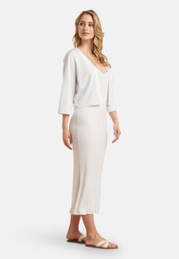 Witte cropped cardigan over een beige geribbelde midi-jurk met een V-hals. Model draagt slide sandalen met een textuur band.