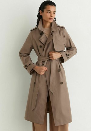 Frau mit dunklen Haaren, die einen beigen Trenchcoat mit Gürtel über passenden Hosen trägt, steht vor einer hellen Wand und blickt zur Seite.