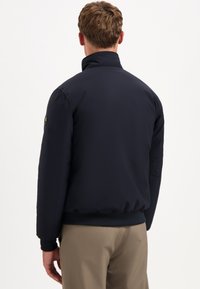 Marineblauwe, gewatteerde jas met hoge kraag en elastische manchetten. Heeft een gladde textuur en een getailleerd ontwerp, gezien vanuit de achterkant.