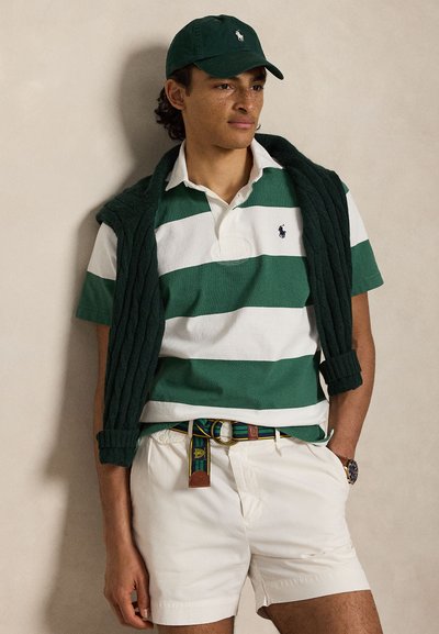 Homme portant un polo rayé vert et blanc, une casquette vert foncé, un pull en maille sombre sur les épaules, un short blanc et une ceinture, les mains dans les poches.