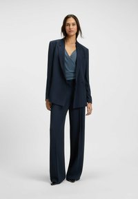 Tummansininen pinstripe-puvusto, jossa on pitkät, leveät housut ja muotoiltu bleiseri, jossa on kärkitaskut, yhdistettynä teksturoituun teal-toppiin.