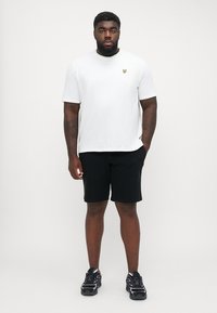 Hvit bomulls T-skjorte med en liten gul logo, kombinert med svarte shorts. Utstyret kompletteres med svarte joggesko.