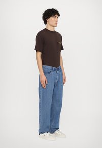 Braunes Baumwoll-T-Shirt mit gelbem Logo, kombiniert mit lässigen blauen Jeans. Das Model trägt weiße Sneakers; sauberes Design, minimalistische Ästhetik.