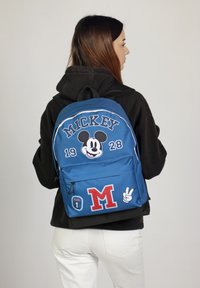 Mochila azul con gráfico de Mickey Mouse, texto "MICKEY 1928" y parches con "M" y "n.º 1". Fabricada en tejido texturizado con correas ajustables.
