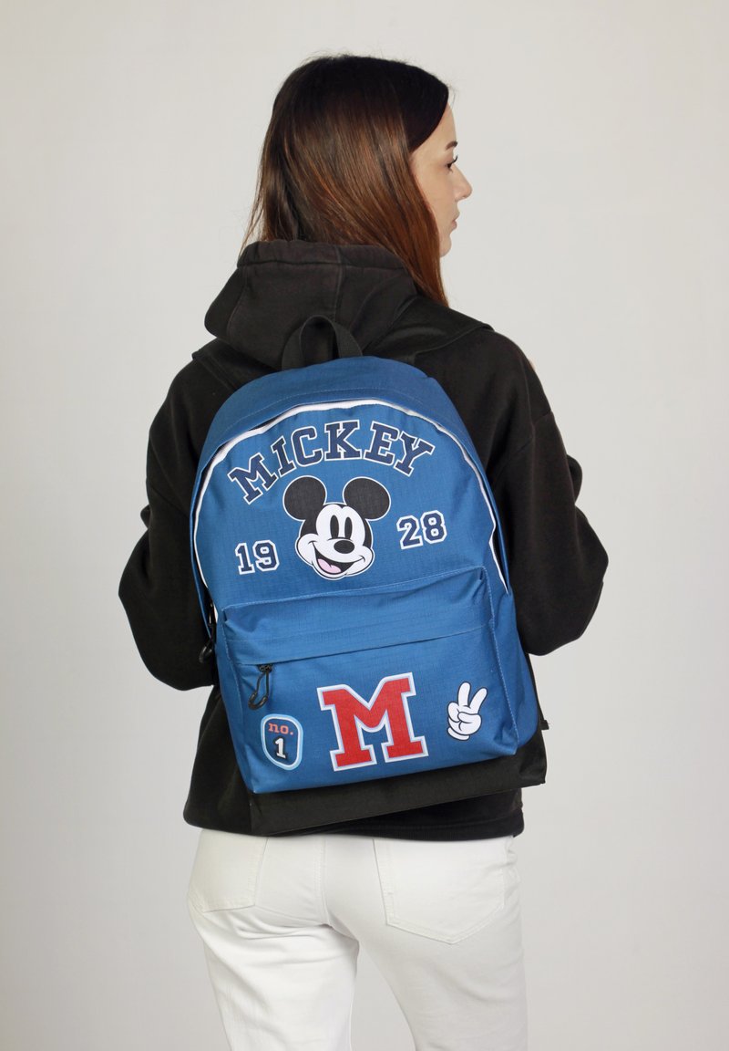 Mochila azul con gráfico de Mickey Mouse, texto "MICKEY 1928" y parches con "M" y "n.º 1". Fabricada en tejido texturizado con correas ajustables.