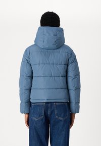 Giacca imbottita con cappuccio di un blu morbido, caratterizzata da cuciture verticali per texture e polsini a coste. Abbinata a jeans di denim blu scuro.