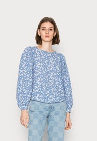 Blusa floral azul clara com mangas longas bufantes e decote redondo, apresentando padrões de flores brancas. Combinada com jeans claros xadrez.