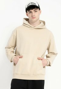 Młody mężczyzna w beżowym hoodie i czarnych spodniach, z rękami w kieszeniach hoodie, w białej i czarnej czapce z napisem "STAY READY."