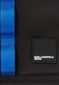 Črna platnena torba z bleščečim modrim trakom in pravokotno črno nalepko z napisom "KARL LAGERFELD JEANS" v beli barvi.