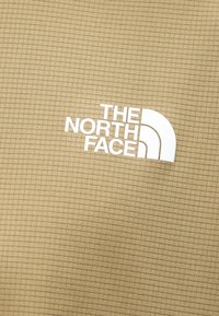 Υφασμα σε μπεζ απόχρωση με υφή, τυπωμένο με το λευκό λογότυπο "THE NORTH FACE" και το σύμβολο του ημικυκλίου.