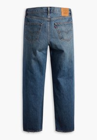 Jean en denim bleu coupe droite avec poches arrière, patch en cuir de marque et coutures visibles sur fond blanc uni.