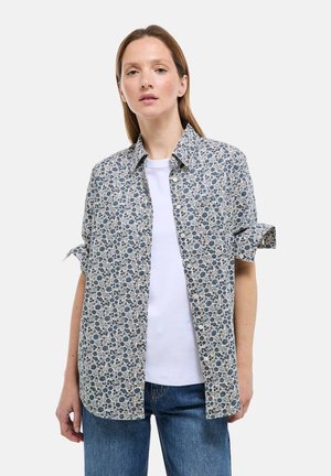 Chemise à motif floral crème avec des fleurs bleues, col classique, manches courtes retroussées, portée par-dessus un t-shirt blanc, jean en denim.