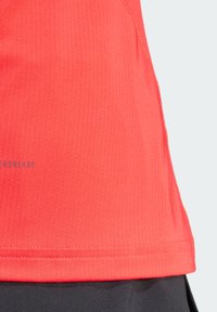 Maglietta atletica leggera di un vivace rosso, con una texture a coste, caratterizzata da un orlo piatto e un piccolo logo in basso. Design a maniche corte.