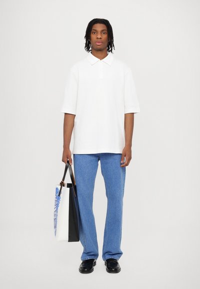White short-sleeve polo shirt, blue denim jeans, black shoes, holding a tote bag with a blue graphic design and black accents.