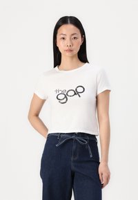 Biely skrátený tričko s čiernym grafickým logom "the gap." V kombinácii s tmavomodrými vysoko pásovými džínami so šnúrkou v páse.