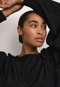 Black, flowing blouse with puff sleeves, featuring gathered detailing at the shoulders. Model's skin tone is a warm beige with natural makeup.