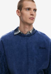 Blue sweatshirt with cable-knit texture on sleeves, featuring a subtle logo. Collar showcases a contrasting patterned shirt underneath.