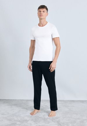 T-shirt bianca in cotone a maniche corte con collo rotondo abbinata a pantaloni neri in tessuto morbido, entrambi senza motivo, indossata da un modello a piedi nudi su uno sfondo chiaro.