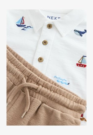 Īsu piedurkņu baltais polo krekls ar zilu un sarkanu izšūtu laivas un vaļa dizainu, papildināts ar brūnām ribotu kokvilnas sporta biksēm ar auklu.