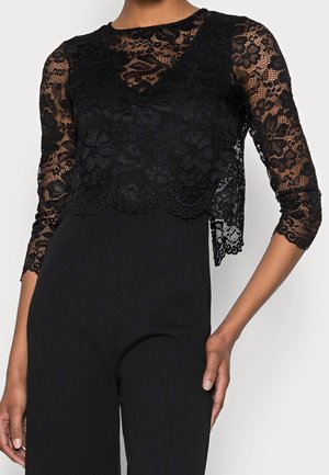 Schwarzes Spitzenoberteil mit Blumenmuster und dreiviertellangen Ärmeln, getragen über einem schlichten, engen schwarzen Kleidungsstück an einer stehenden Person.