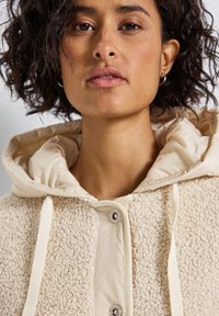 Gros plan sur une femme aux cheveux bouclés et foncés, portant une veste texturée couleur crème avec capuche et cordons de serrage.
