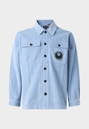 Jeune homme aux cheveux bouclés portant une veste en velours côtelé bleu clair avec des boutons noirs et un écusson 8-ball sur la poche poitrine.