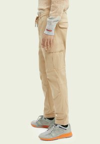 Person iført beige cargo bukser, grå og orange sneakers, samt en lys beige sweater, der står mod en ensfarvet baggrund.