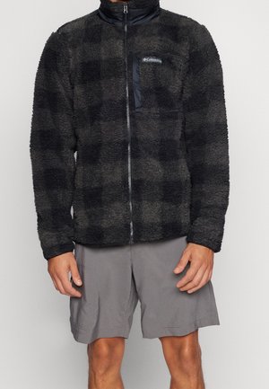 Mężczyzna ubrany w czarno-ciemnoszarą flanelową kurtkę zapinaną na zamek, w połączeniu z szarymi spodenkami do kolan, stojący na jednolitym tle.