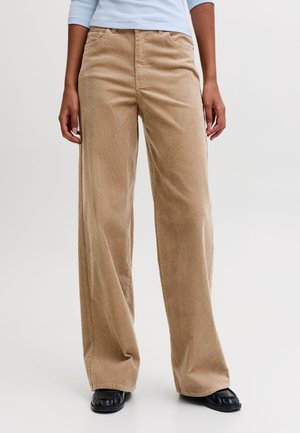 Personne portant un pantalon large en velours côtelé beige, un haut bleu clair et des chaussures noires, debout devant un fond uni.