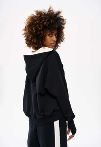 Zwarte oversized hoodie met een witte kraag, voorzien van een kangoeroezak en geribde manchetten. Model poseert achterwaarts en toont het ontwerp aan de achterkant.