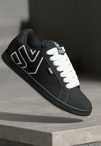 Schwarze und graue Sneaker mit weißen Schnürsenkeln und einem auffälligen weißen Chevron-Design, die über einer Steinoberfläche schwebend präsentiert werden.