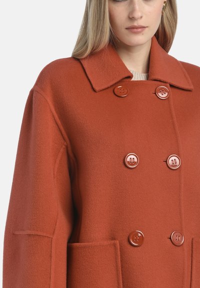 Palton dublu, într-o culoare bogată de rugină, confecționat din lână texturată. Dispune de butoane mari, lucioase și un guler cu tăietură. Include buzunare laterale.