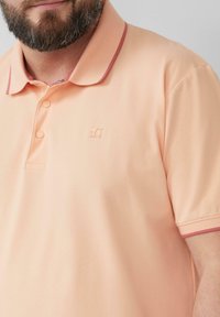 Pfirsichfarbene Poloshirt aus strukturiertem Stoff, mit kontrastierenden mahagonifarbenen Akzenten am Kragen und an den Ärmeln sowie einer Zweiknopf-Leiste. Verfügt über ein kleines Logo.