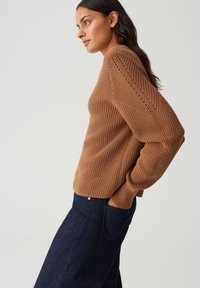 Frau in einem braunen Strickpullover mit strukturierten Maschen, kombiniert mit dunkelblauen hochgeschnittenen Jeans, die im Profil steht und die Hände in den Taschen hat.