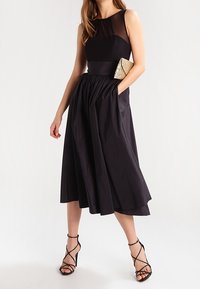 Abito nero senza maniche con un corpino aderente e una gonna svasata. Pannello trasparente sul collo. Gli accessori includono una pochette dorata e tacchi con cinturino.