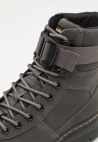 Botas de tobillo de ante gris con un cuello acolchado, que presentan herrajes negros, ojales hexagonales y costuras a contraste. Correa ajustable para un mejor ajuste.