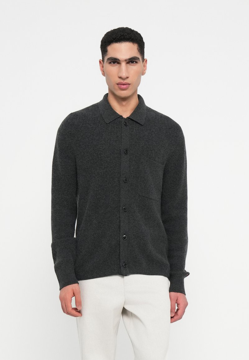 Sötét szürke kötött kardigán, gallérral, gombos elülső résszel, hosszú ujjakkal és mellzsebbel. Világos színű nadrág fölé van hordva.