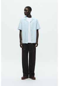 Chemise bleu clair à manches courtes avec col et fermeture à boutons, portée sur un t-shirt blanc ; associée à un pantalon large noir et des chaussures brunes.