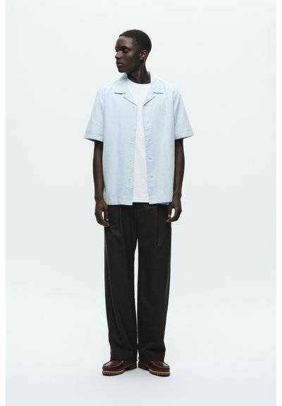 Chemise bleu clair à manches courtes avec col et fermeture à boutons, portée sur un t-shirt blanc ; associée à un pantalon large noir et des chaussures brunes.