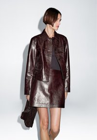 Frau trägt eine dunkelburgunderrote Lederjacke und passenden Rock, hält eine dunkel Wildledertasche und schaut seitlich vor einem einfarbigen Hintergrund.