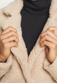 Beige, getextureerde nepbontjas met grote revers, gedragen over een zwarte coltrui. Handen grijpen de kraag vast, waarbij de zachtheid van de stof wordt benadrukt.