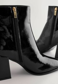 Czarne lakierowane botki za kostkę z wydłużonym noskiem, widocznymi suwakami na bokach i masywnymi kwadratowymi obcasami. Charakteryzują się gładkim, błyszczącym wykończeniem.