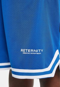 Bermudas deportivas azules de tejido texturizado, con una franja blanca en el dobladillo y el logotipo impreso "DEPARTAMENTO CREATIVO RETERNITY" cerca de la parte inferior.
