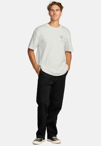 Light gray, short-sleeve t-shirt with a palm tree graphic on the left chest, paired with black pants and black sneakers.