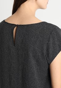 Czarna bluzka w białe kropki, z okrągłym dekoltem, krótkimi rękawami i zapięciem na plecach w formie łezki z ozdobnym guzikiem. Gładka struktura tkaniny.