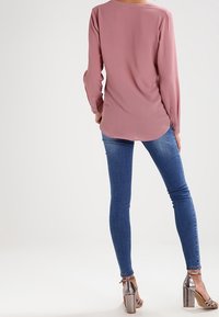 Blouse rose pâle à manches longues avec un ourlet arrondi, associée à un jean bleu ajusté et des sandales à talons hauts argentées, mettant en valeur une texture lisse.