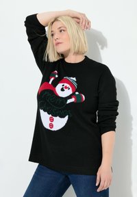 Blonde Frau in schwarzem Weihnachtspullover mit Schneemann und dem Text "Frohe Weihnachten", posiert mit einem erhobenen Arm vor einem weißen Hintergrund.