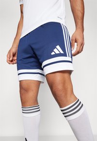 Marina atletiska shorts med vita accenter, med tre vita ränder och en Adidas-logotyp. Kombinerat med knähöga vita strumpor med svarta ränder.