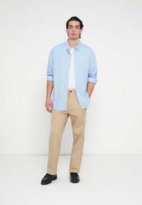 Πουκάμισο με κουμπιά με ανοιχτό μπλε και λευκές ρίγες, φορεμένο πάνω από λευκό T-shirt, με μπεζ παντελόνι και μαύρα παπούτσια, στέκεται μπροστά σε απλό φόντο.